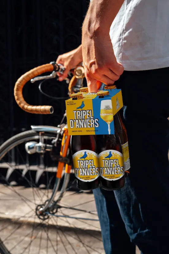 Foto van een man, met in de ene hand een pakket Tripel D'Anvers bier en de andere hand een oude koersfiets.